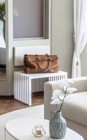 A modern living room with a white sofa, a round coffee table with a vase and candle, a bench with a brown leather bag, and a window with curtains.