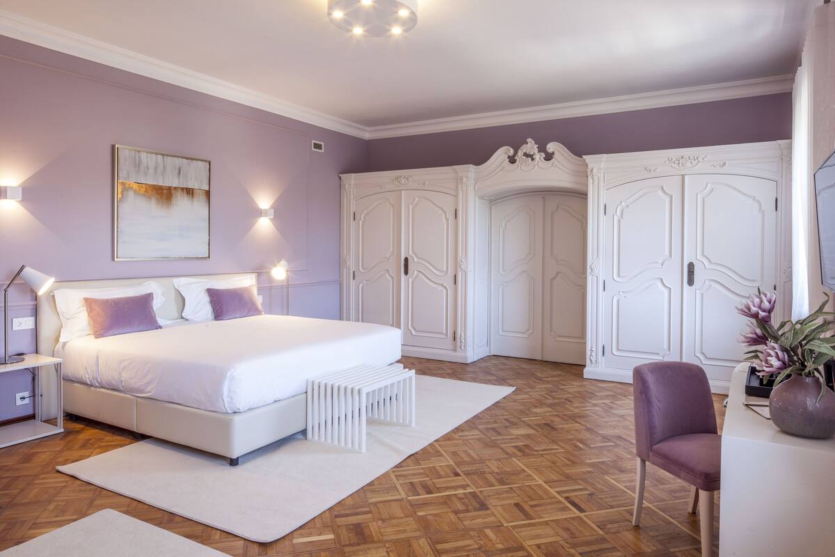 A bedroom with a large bed, white wardrobe, and a desk with a chair.