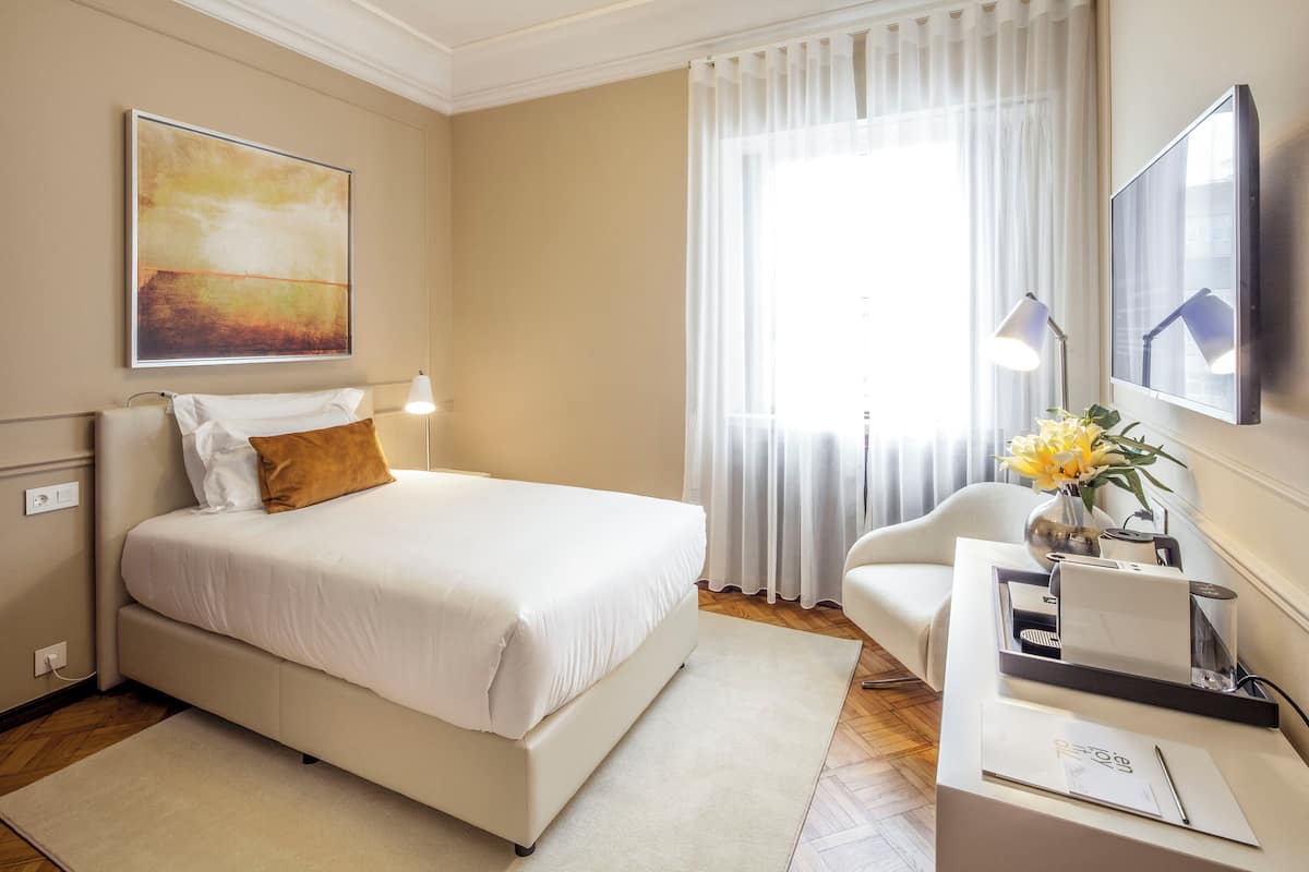 A hotel room with a bed, a desk with a lamp, a chair, a vase with flowers, and a painting on the wall.