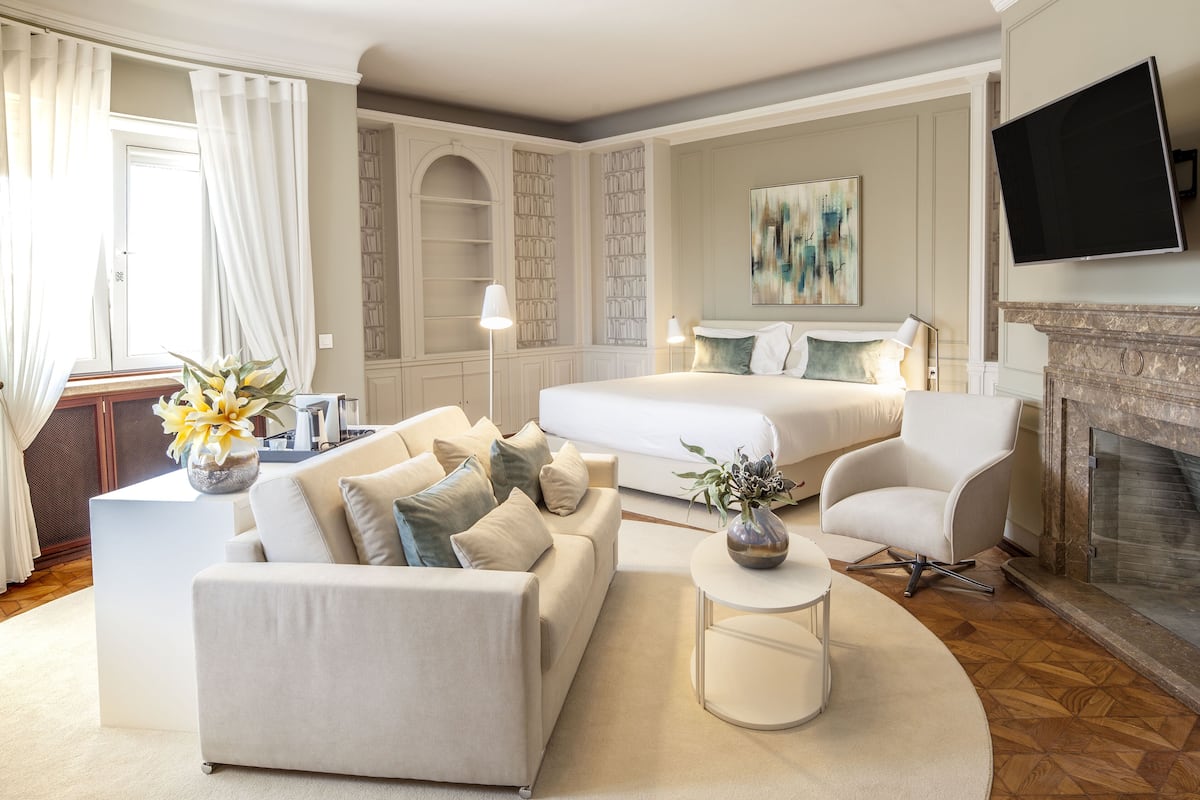 A modern hotel room with a large bed, a sofa, a small table, and a fireplace.