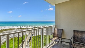 A balcony with wicker furniture overlooking a beach with umbrellas. - Destin On The Gulf 406 2 Bedroom Condo by RedAwning (Destin)
