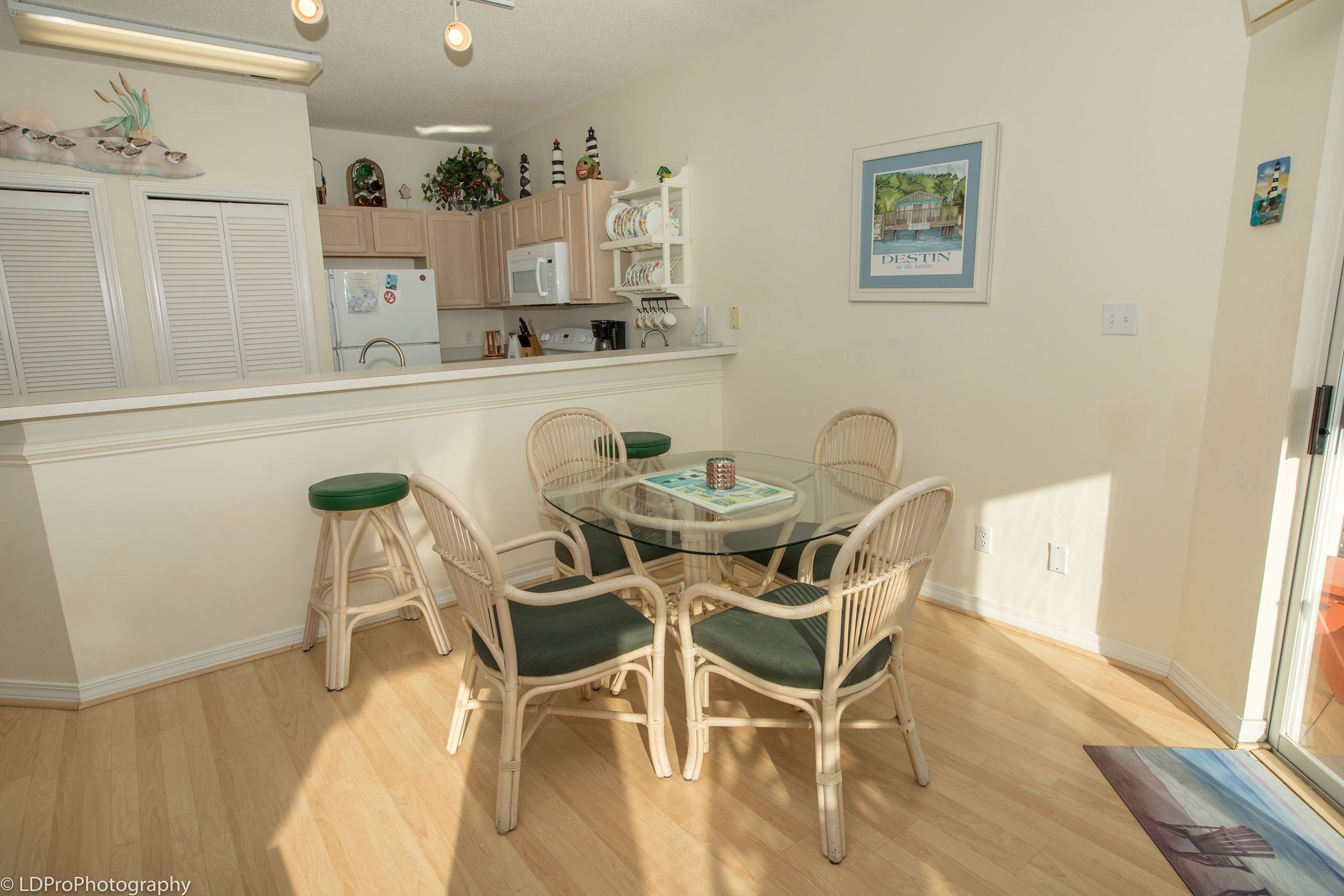A dining area with a glass-top table surrounded by wicker chairs with green cushions, a kitchen in the background, and a framed picture on the wall.