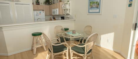 A dining area with a glass-top table surrounded by wicker chairs with green cushions, a kitchen in the background, and a framed picture on the wall.