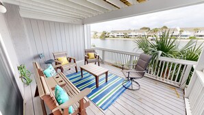 A covered outdoor deck with wooden furniture, a striped rug, and a view of a body of water and buildings in the background. - South Bay By The Gulf 062 2 Bedroom Townhouse by RedAwning (Destin)