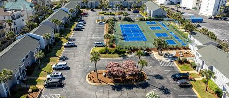 An aerial view of a parking lot with a tennis court, surrounded by residential buildings and a beach.