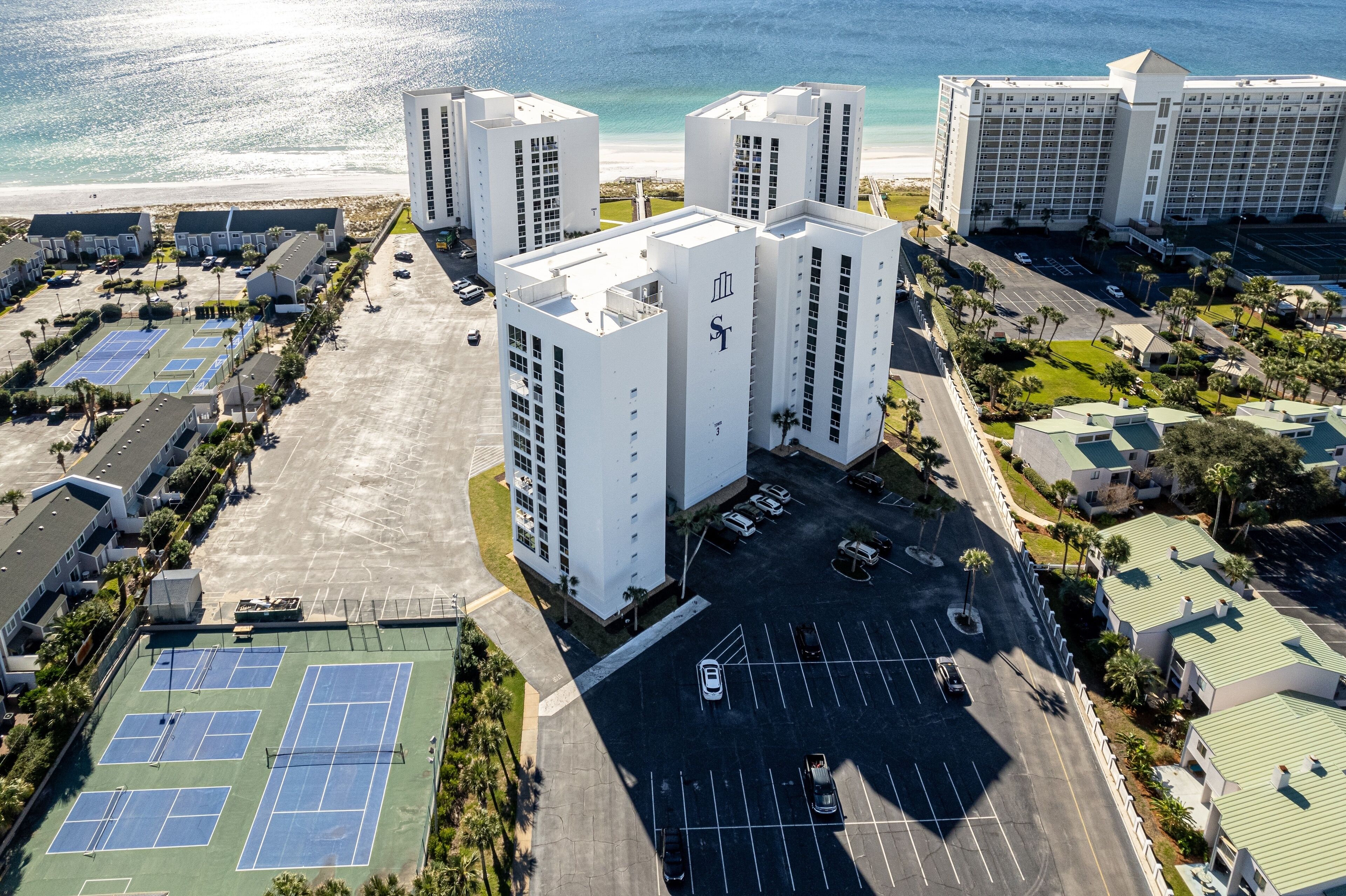 Vista aérea de un complejo hotelero con varios edificios altos, un estacionamiento y canchas de tenis.