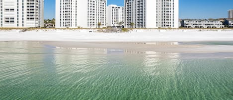A beachfront hotel with multiple high-rise buildings, clear shallow waters, and a sandy beach.