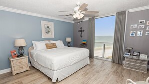 Un dormitorio con cama, ventilador de techo, mesita de noche con lámpara, pequeña mesa con lámpara y vista a la playa a través de un ventanal grande.