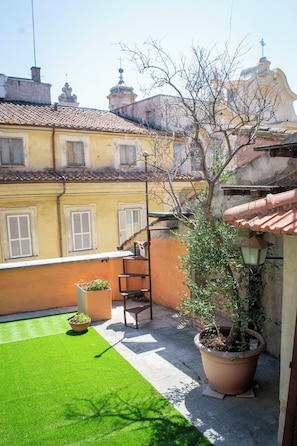 A courtyard with a green lawn, a potted plant, a metal chair, and a building with yellow walls and windows. - Charme & Chic Luxury (Rome)