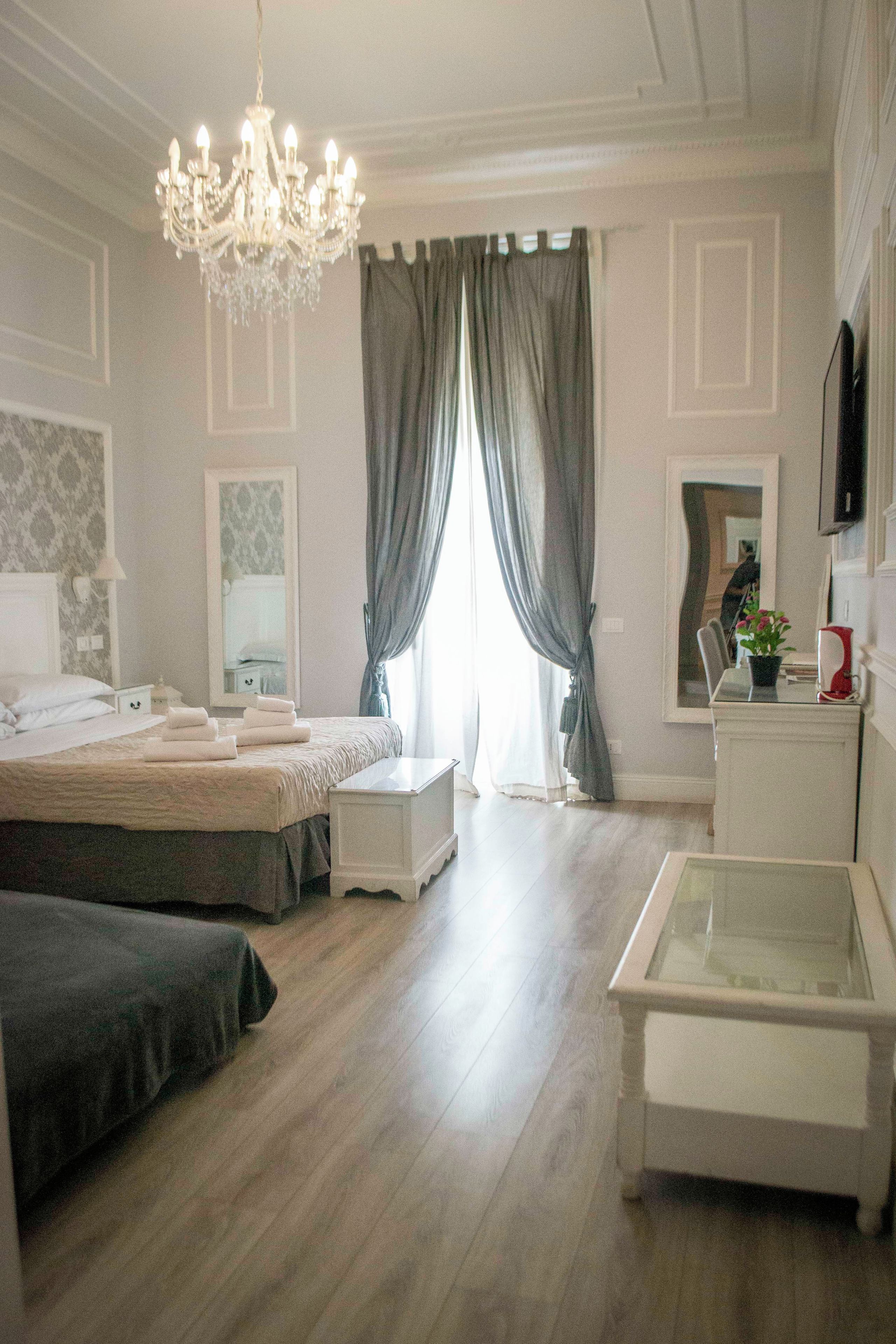 A bedroom with a bed, bedside tables, a mirror, a chandelier, a window with curtains, and a television on the wall.