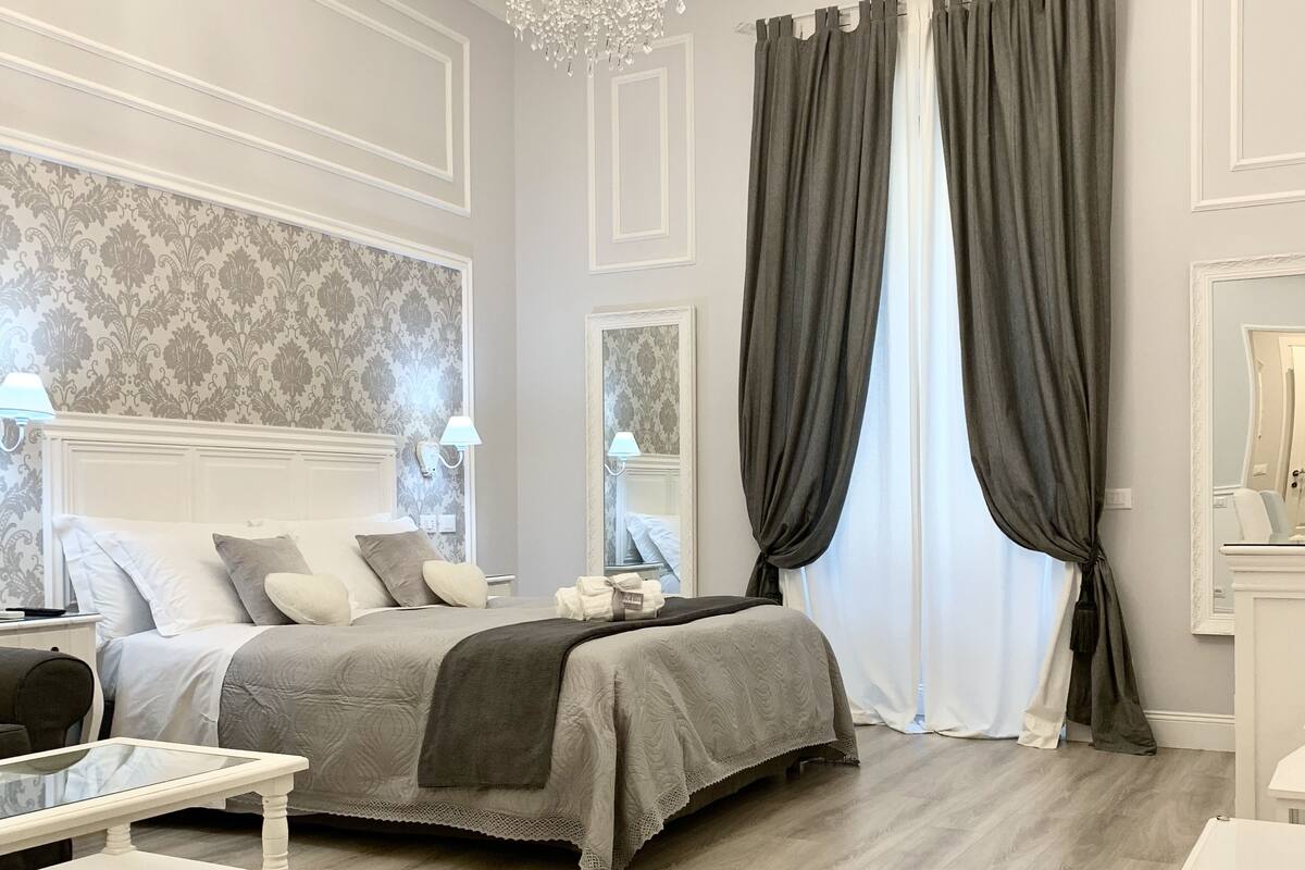 A bedroom with a bed, bedside tables, a chandelier, and a large window with curtains.