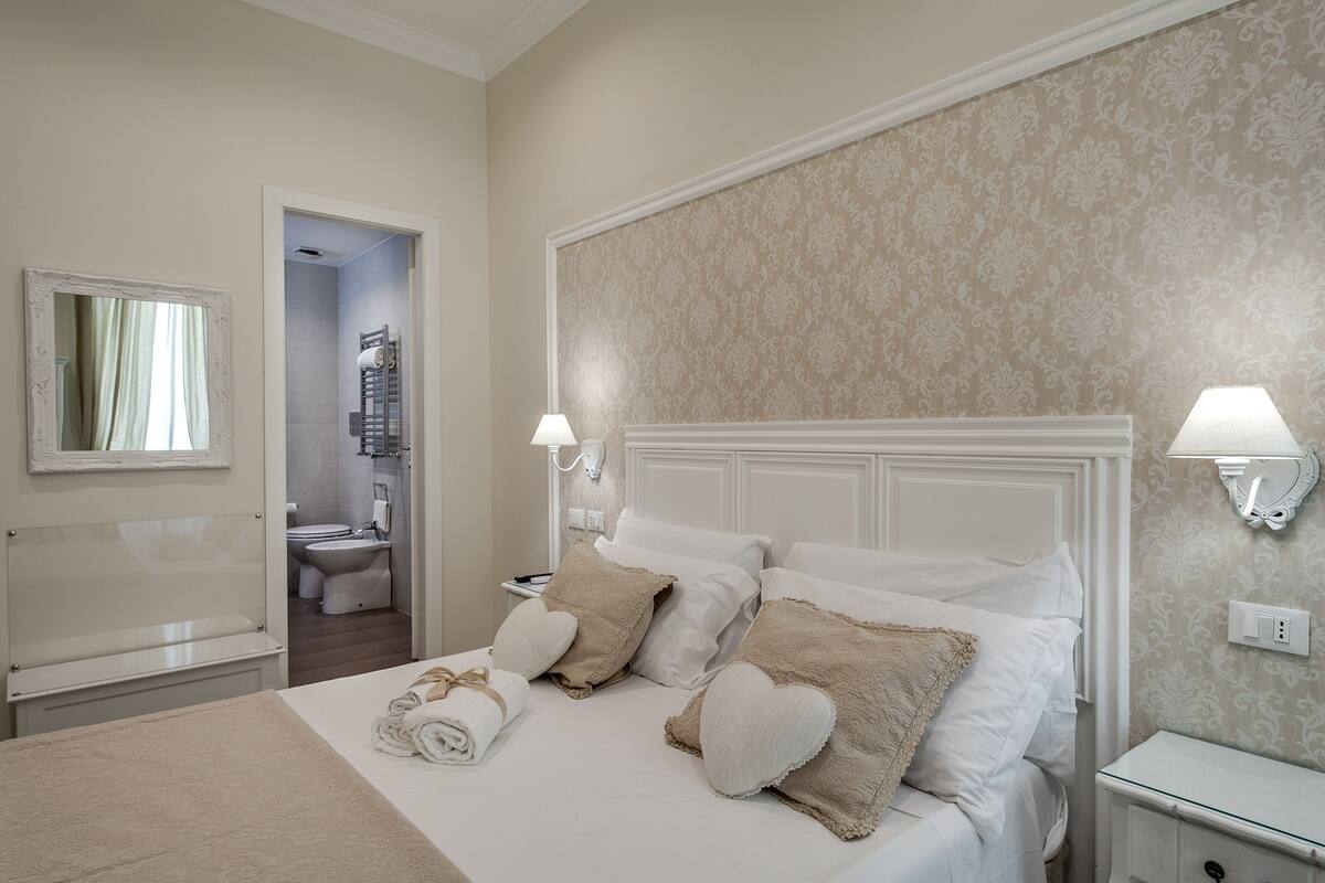 A hotel room with a bed, two bedside lamps, a nightstand, and a bathroom visible through an open door.