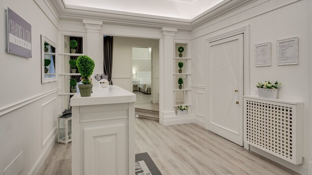 Un pasillo blanco con una recepción, plantas en macetas y una puerta que da a otra habitación.