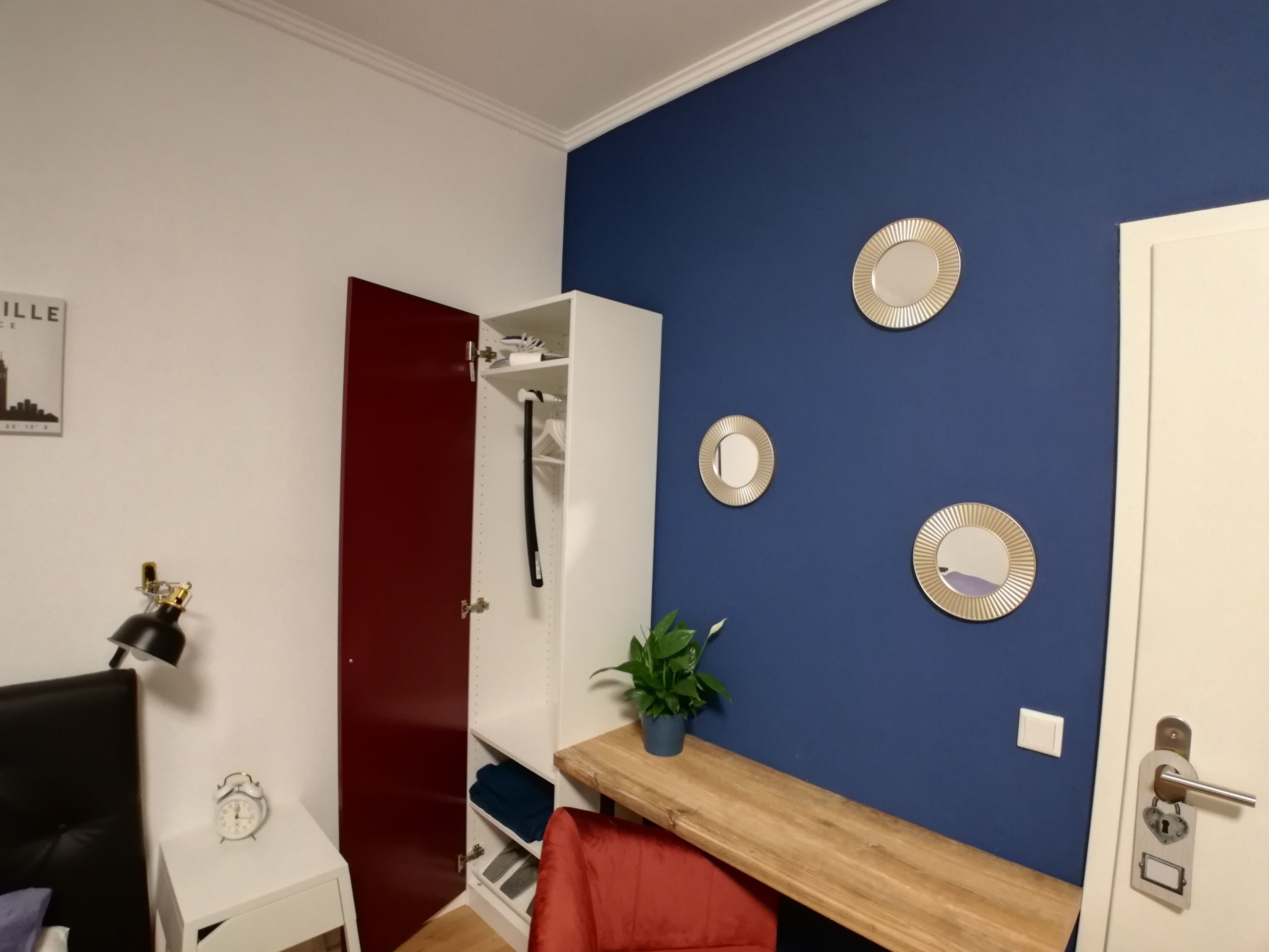 A room with a red door, a white wall with a cityscape poster, a wooden desk with a plant, and a blue wall with decorative plates.