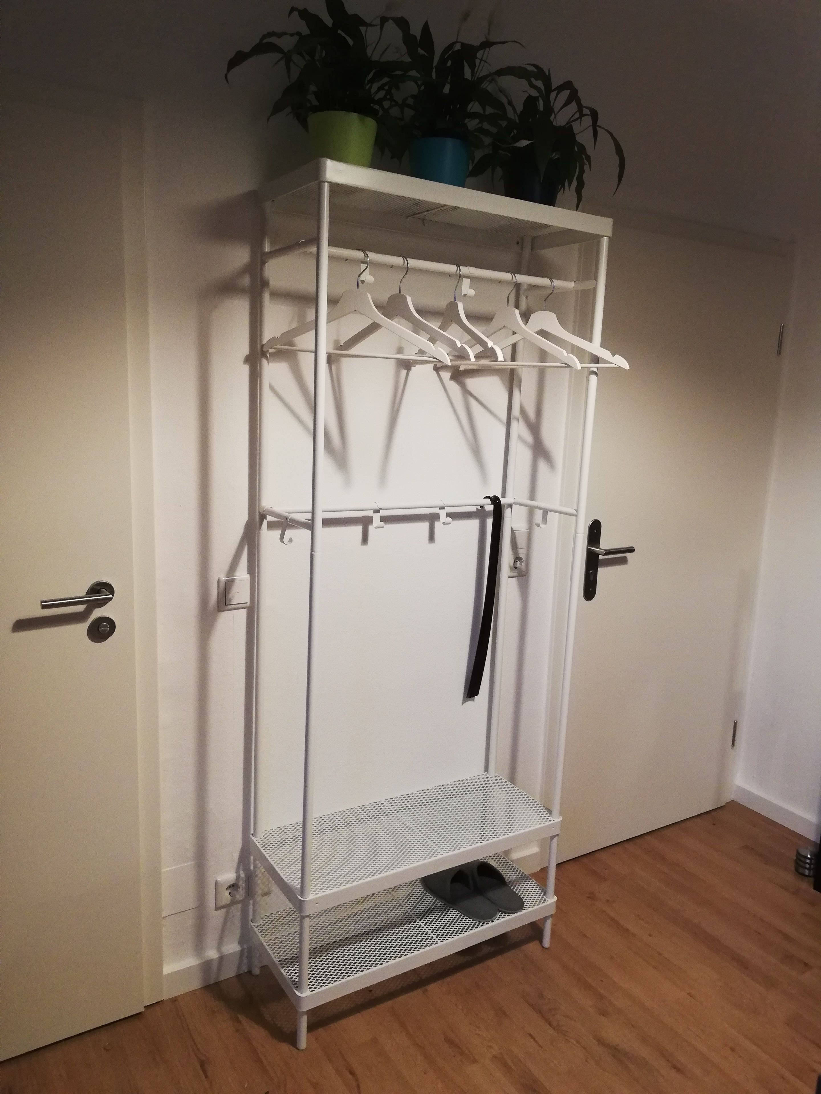 A white metal wardrobe with shelves and a hanging section, a pair of black shoes on the bottom shelf, and a potted plant on top.