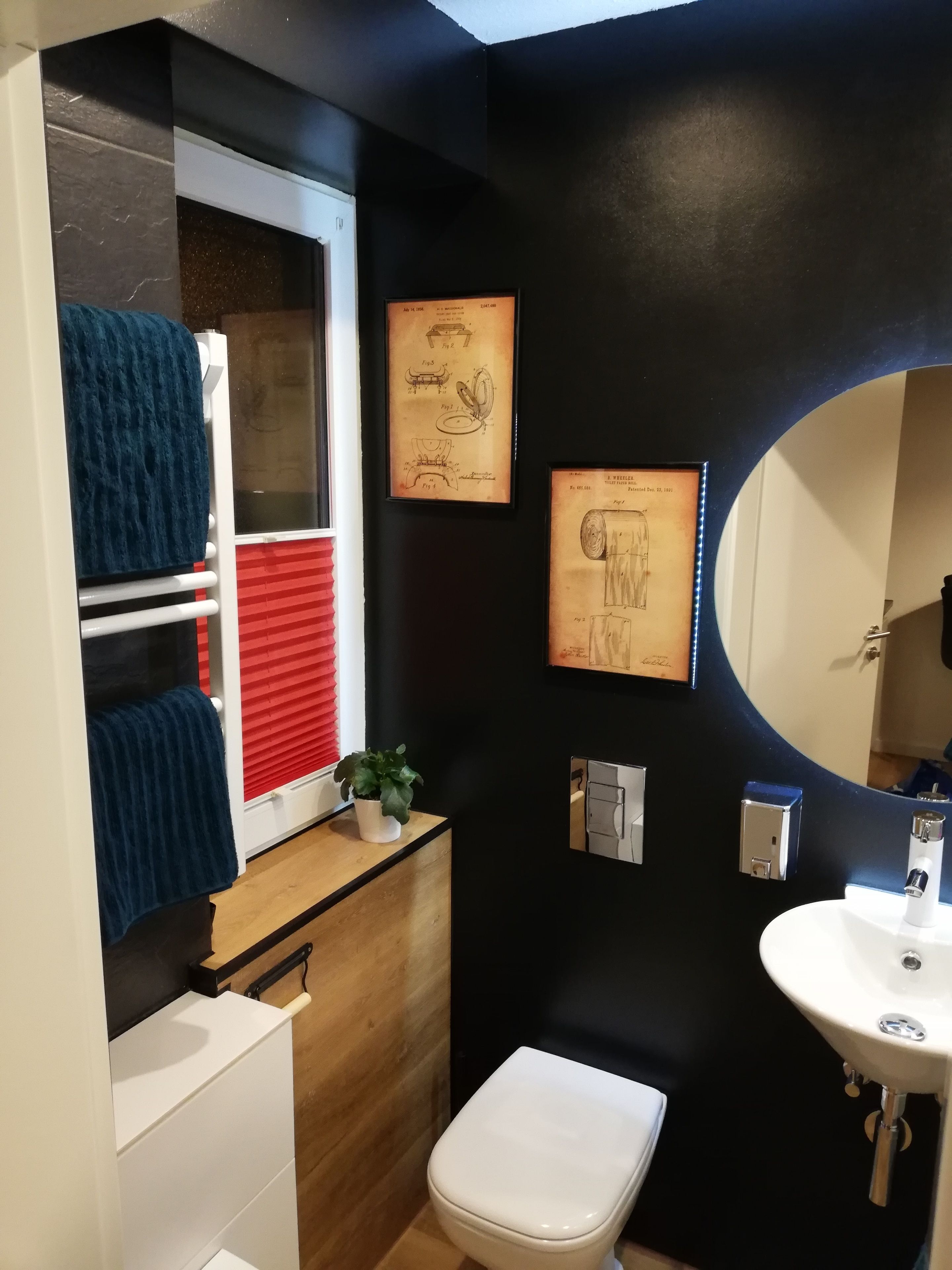 A compact bathroom with a white toilet, a sink with a mirror, and a window with red blinds. There are towels hanging on a rack and a small potted plant on the windowsill.
