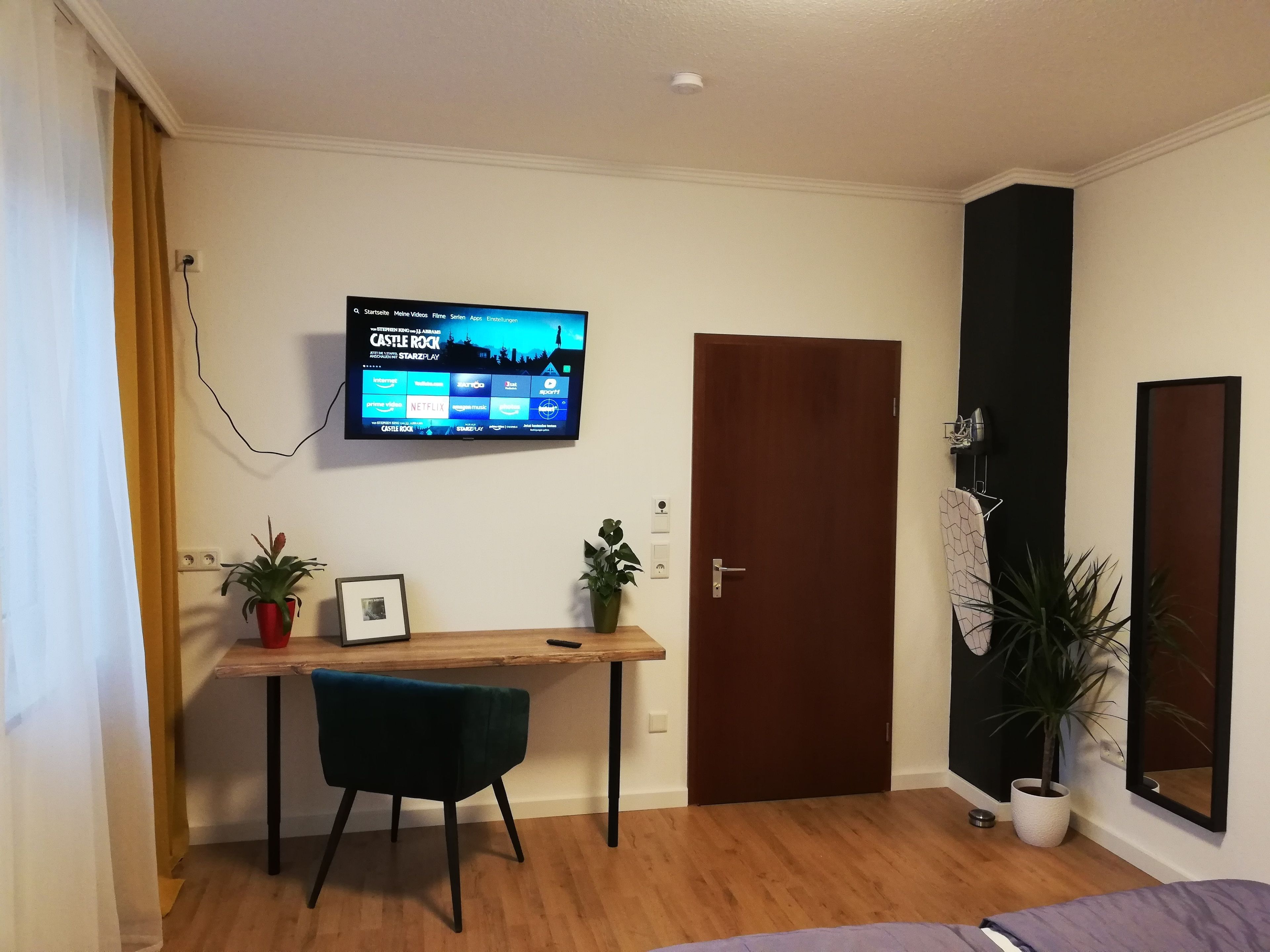 Ett rum med ett skrivbord i trä, en stol, en tv som är monterad på väggen, en dörr och en krukväxt.