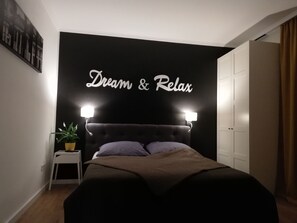 A bedroom with a black headboard, two bedside lamps, a bed with a brown blanket, a small bedside table with a plant, and a wall with the words "Dream & Relax". - Dream & Relax Apartment's Messe (Nuremberg)