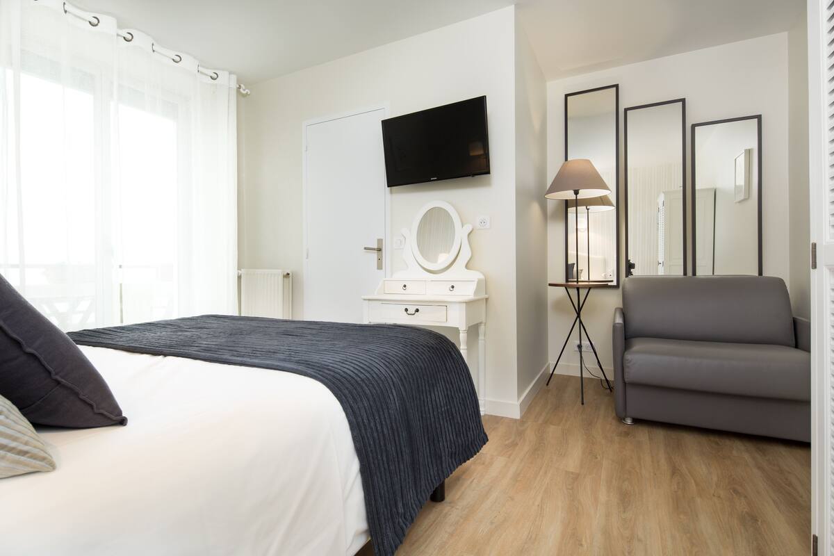 Un dormitorio con cama, mesita de noche, espejo, televisor y un sillón gris.