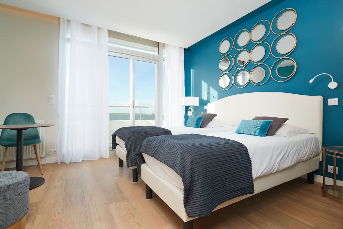 Una habitación de hotel moderna con una cama grande, una zona de estar con una mesa y sillas, y vistas al exterior.
