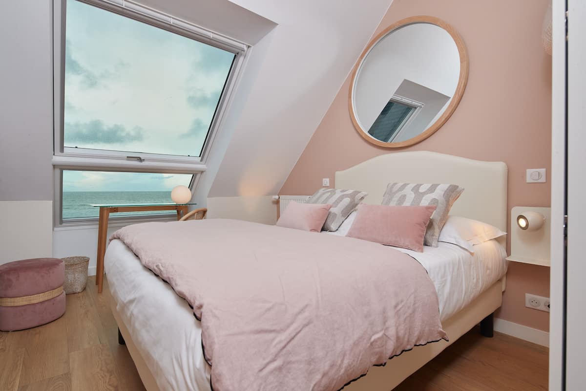 Un dormitorio con una cama grande, un espejo redondo, una mesita de noche y vistas al mar a través de una ventana.