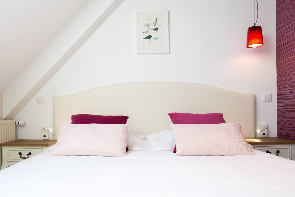 Un dormitorio con cama blanca, dos almohadas, una mesita de noche, una lámpara colgante roja, un radiador e ilustración enmarcada de un pájaro en la pared.