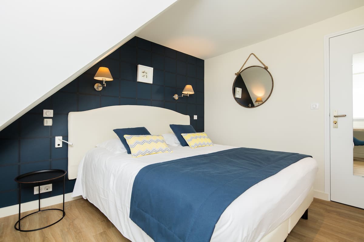 Un dormitorio con una cama blanca, almohadas con estampado azul y amarillo, una colcha azul, un espejo redondo y dos lámparas de pared.