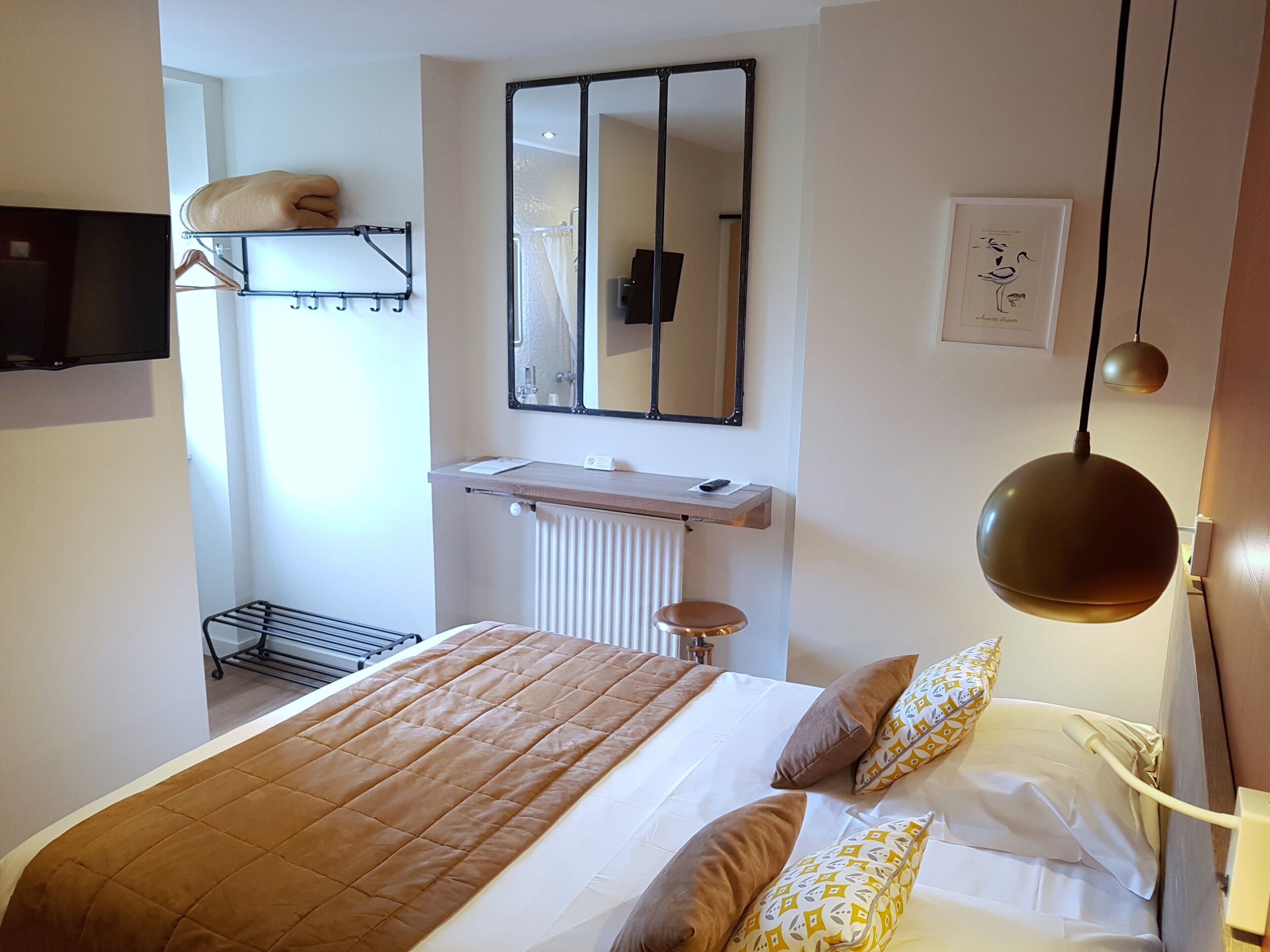 A hotel room with a bed, a bedside table, a mirror, a shelf with towels, and a TV.