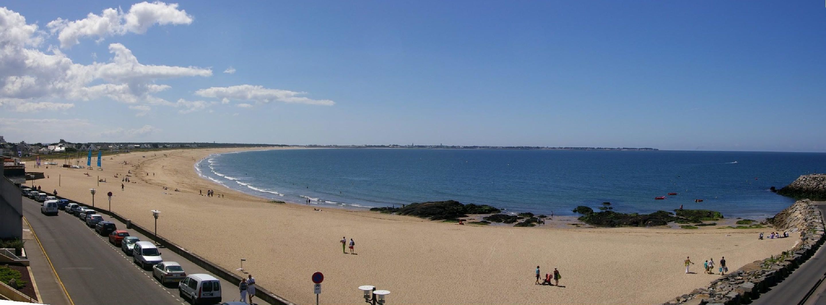 Une plage bordée d’une route, quelques personnes qui se promènent et un ciel bleu dégagé.