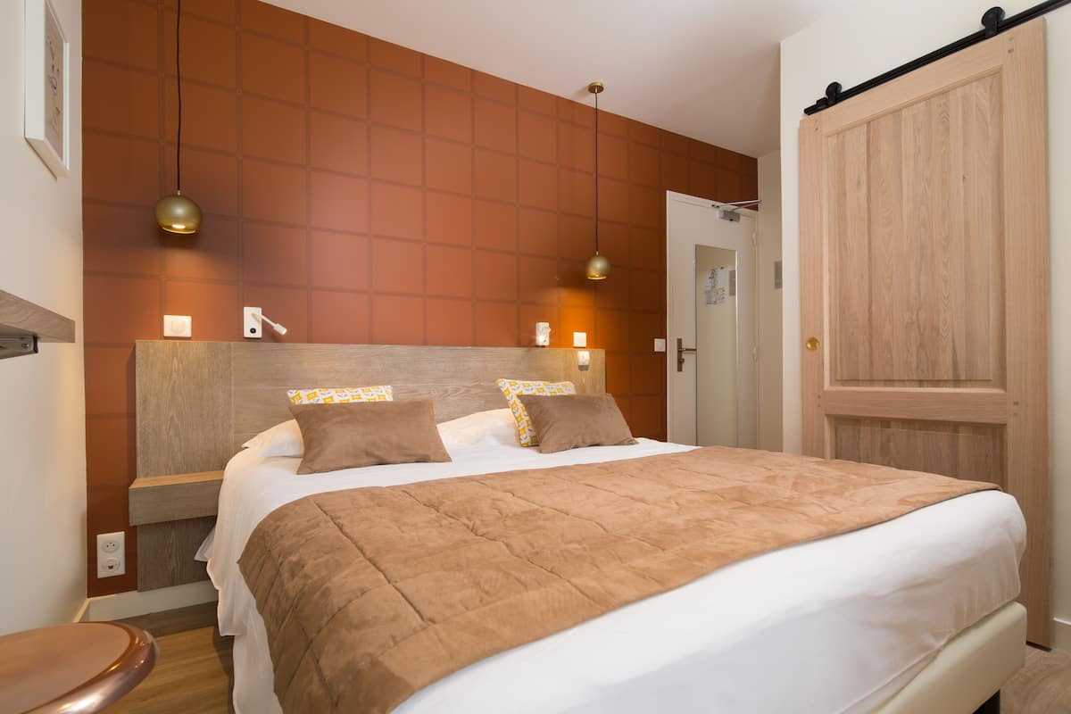 Una cama bien hecha con una manta y almohadas marrones, un cabecero de madera y dos lámparas colgantes.