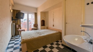 A hotel room with a checkered floor, a bed, a television mounted on the wall, a bathroom with a sink, and a view of buildings through the window. - Vitalo 402 (Koksijde)