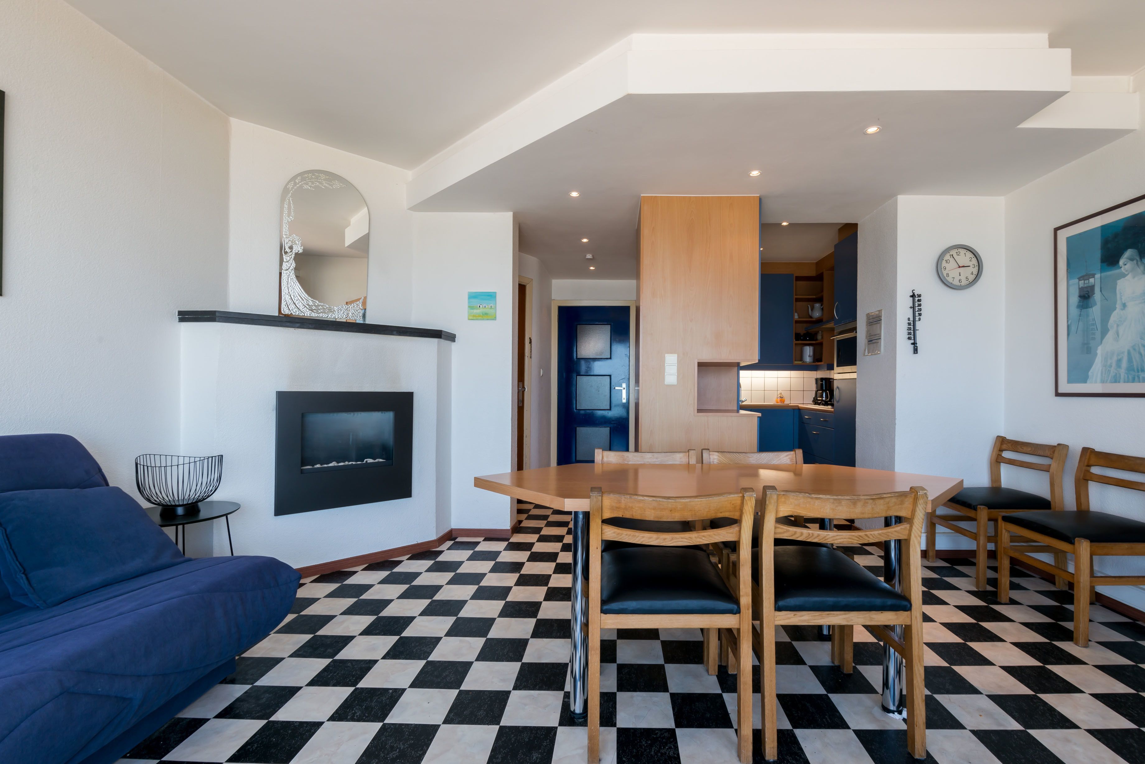 Un salon moderne avec un sol à carreaux, une cheminée, une table à manger avec des chaises, un canapé et un coin cuisine visible à travers une porte ouverte.