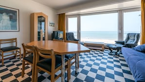 A room with a checkered floor, a wooden table with chairs, a sofa, a television, and a large window with a view of the sea. - Vitalo 402 (Koksijde)
