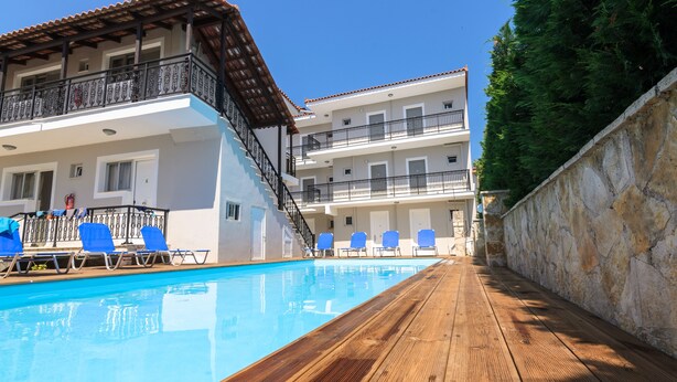A swimming pool with blue chairs, a wooden deck, and a building with balconies and windows. - Karras Livin (Zakynthos)