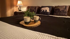 Ein Bett mit dunkler Bettdecke, zwei Kaffeetassen auf einem Holzuntersetzer, eine Topfpflanze und eine Lampe in einem Zimmer mit einem gerahmten Bild und dekorativen Kissen.