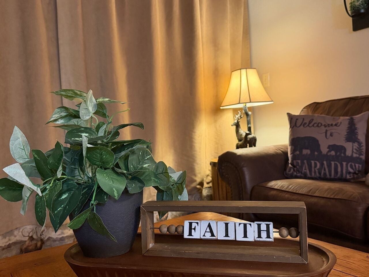 A potted plant, a wooden tray with the word 'FAITH' spelled out, and a lamp on a table in a room with a brown leather couch and a cushion with a bear design.