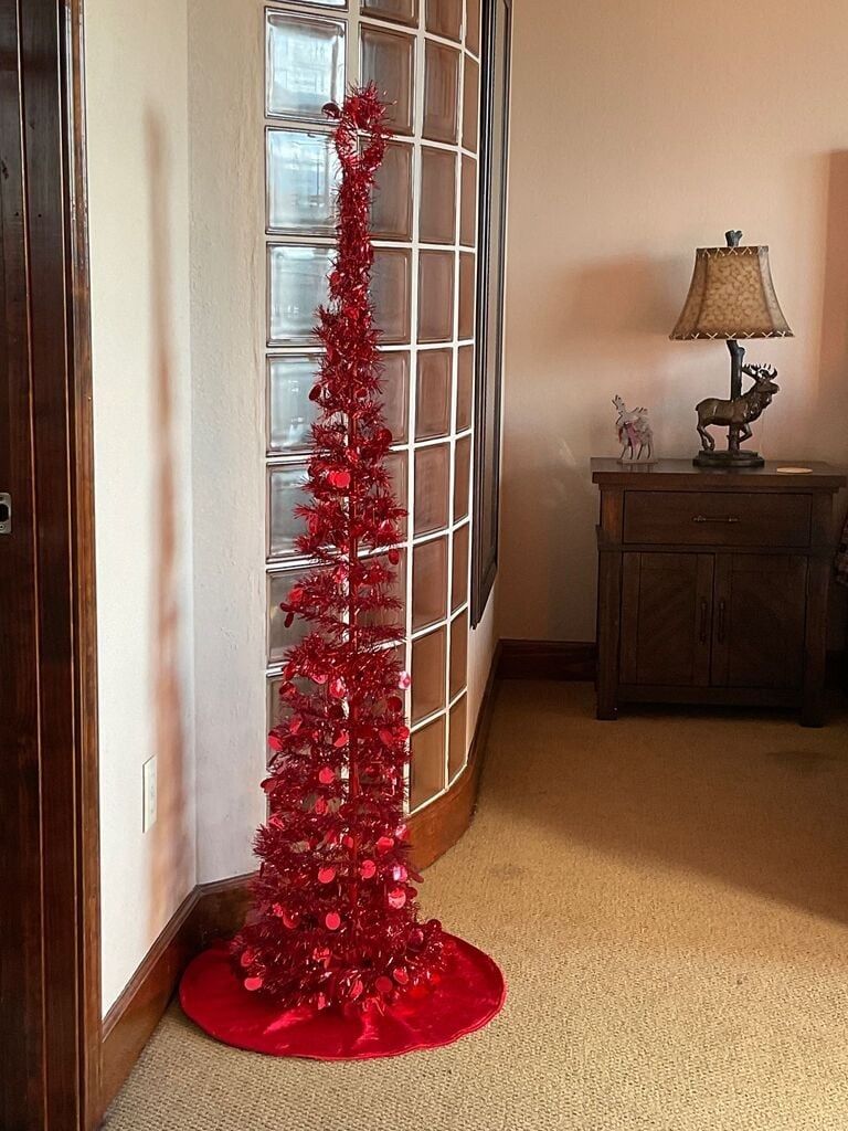 A red Christmas tree in a corner with a lamp and a cabinet in the background.