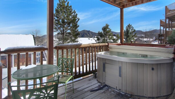 Une terrasse en bois avec un jacuzzi, une table en verre et une chaise, offrant une vue sur un paysage enneigé et montagneux.