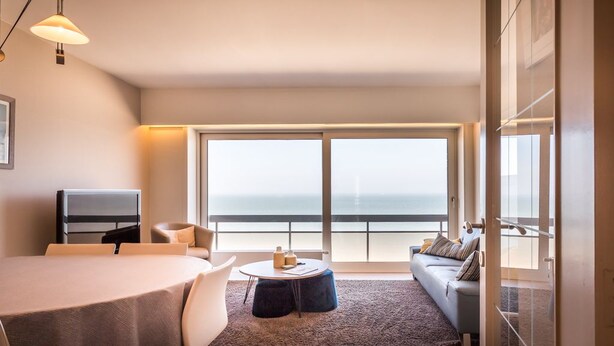 A modern living room with a large window, a round dining table, a sofa, and a TV. - Kentucky 603 (Koksijde)