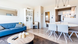 A modern living room with a blue sofa, a round coffee table, and a dining area with white chairs and a table. - Kentucky 603 (Koksijde)