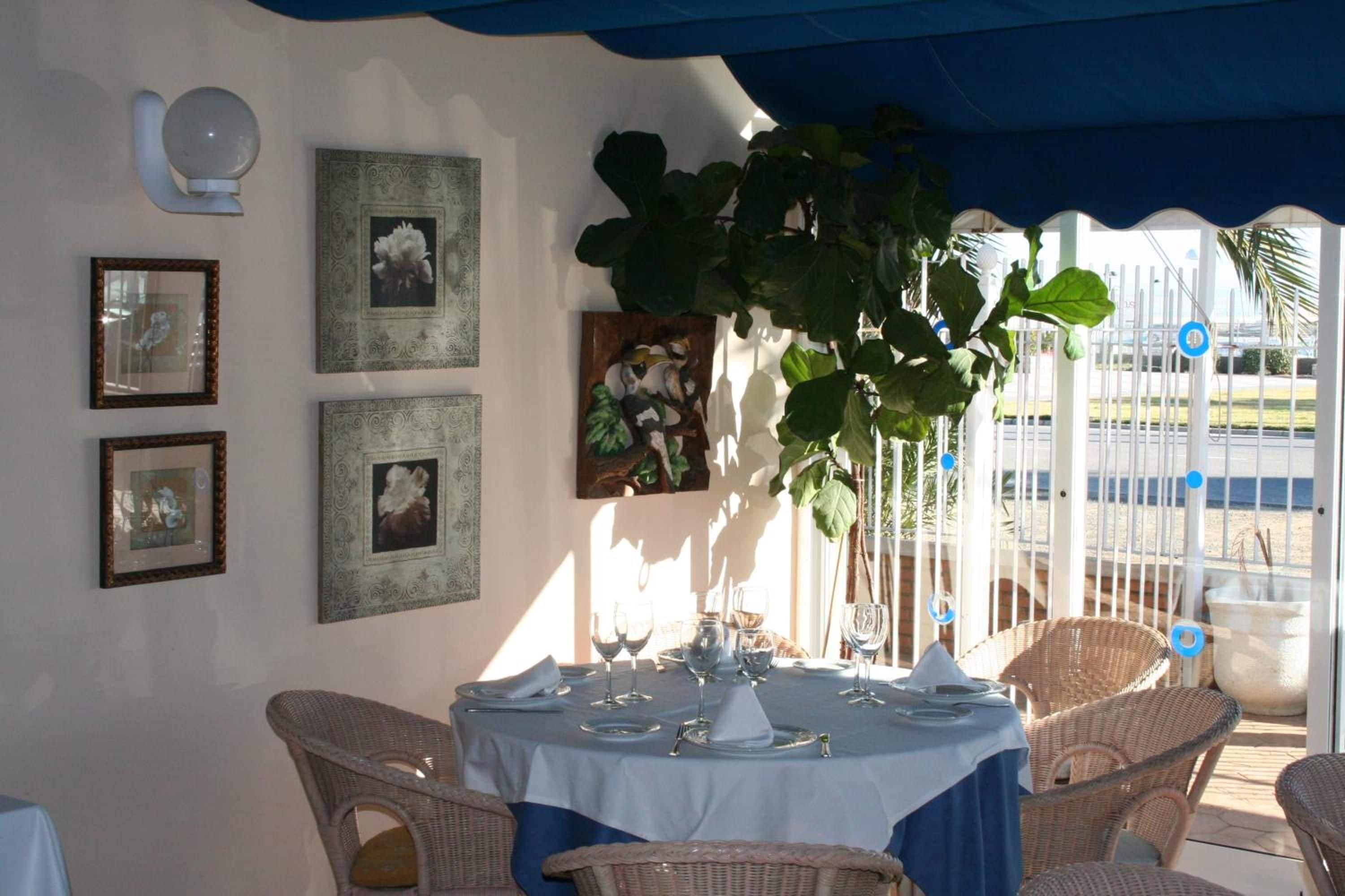 A dining area with wicker chairs, a round table set with plates, glasses, and cutlery. There are framed pictures on the wall and a large plant hanging above the table.