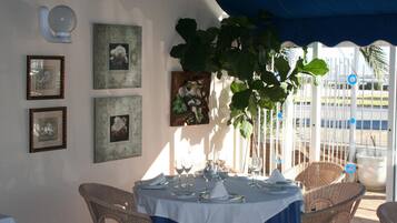 A dining area with wicker chairs, a round table set with plates, glasses, and cutlery. There are framed pictures on the wall and a large plant hanging above the table.