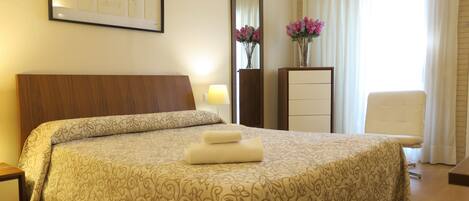 Habitación de hotel con una cama grande, una mesita de noche, un aparador, un espejo, una lámpara, una silla y un florero con flores.
