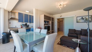 A modern kitchen with blue cabinets, a dining area with a glass table, and a living room with a brown sofa and a glass coffee table. - San Remo 501 (Koksijde)