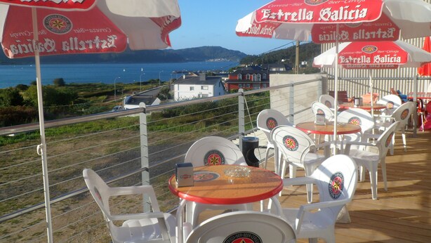 Outdoor seating with red umbrellas, white chairs, and a view of the sea and buildings. - Hospedaxe Don Manuel (O Vicedo)