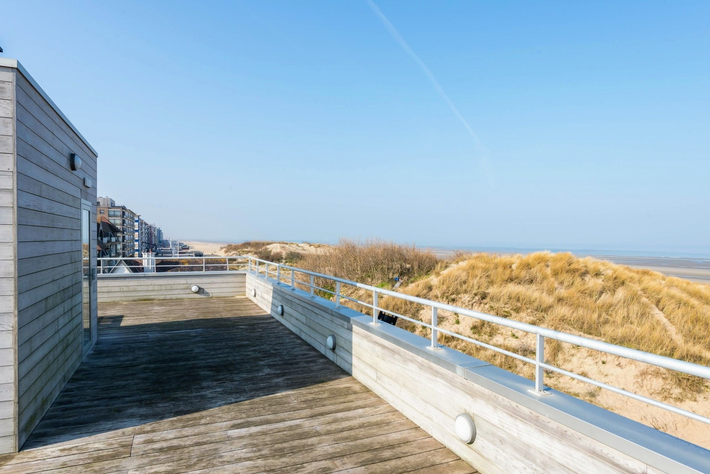 Une terrasse en bois avec une rambarde donnant sur une plage et des dunes.