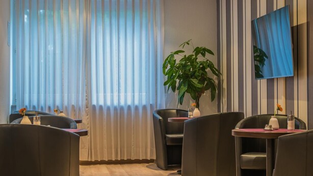 A modern dining area with black leather chairs, a table with a vase of flowers, and a large window with sheer curtains. - Hotel am Wasen (Freiberg am Neckar)
