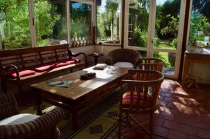 Una habitación iluminada por el sol con una mesa de madera, sillas de mimbre y un banco con cojines rojos.