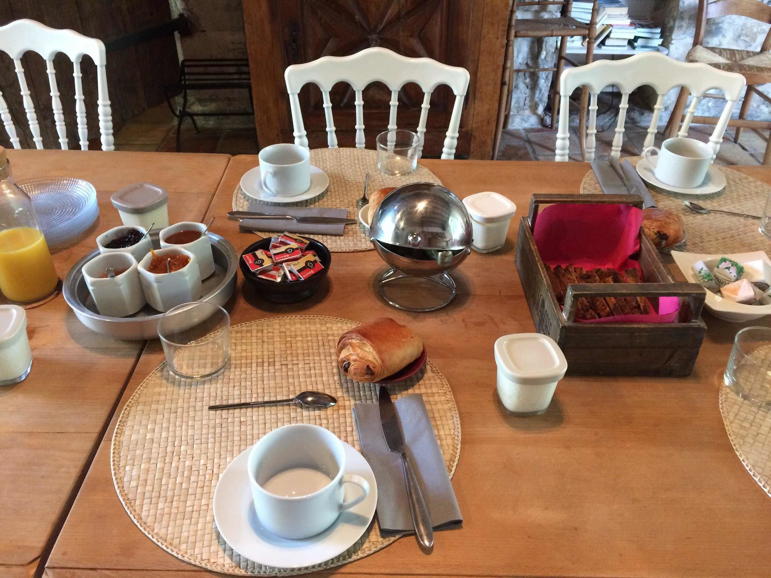 A breakfast setting with a table set for two, including plates, cups, a bowl, and a basket of bread.