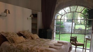 A bedroom with a large bed, a view of the garden through a window, and a chair by the window. - Maison Basta (Orthevielle)